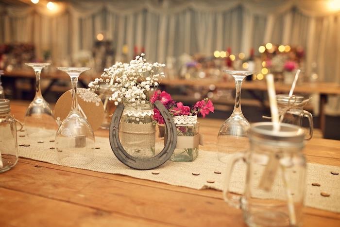 kaimiškas kaimiškas vestuvių stalo dekoras, džiuto stalo bėgikas, pasagos centras, nėriniais dekoruoti stikliniai indeliai ir džiuto su lauko gėlėmis