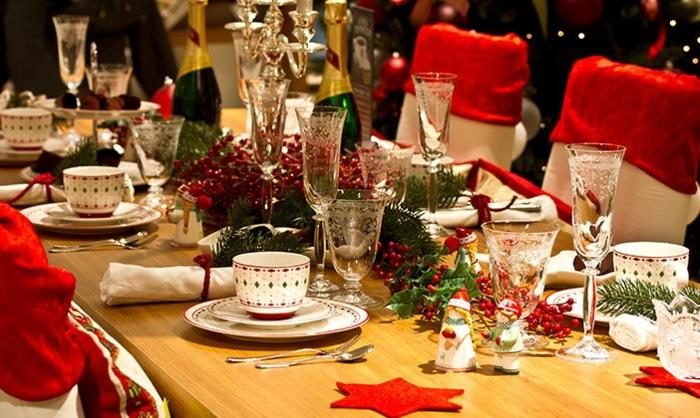 ilgas medinis stalas, baltos lėkštės ir spalvoti raštuoti kavos puodeliai, raudonos žvaigždės, vyno taurės, žalias dekoras, butelis šampano