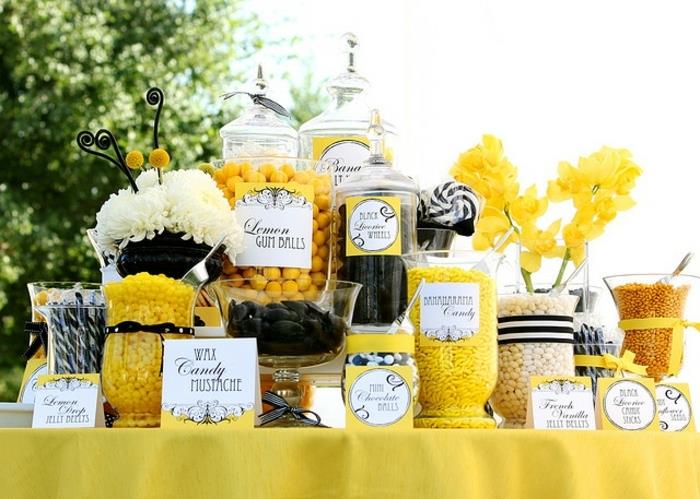 sladkani mandlji in lizike v črni, beli in rumeni barvi, rumeni prt, nalepke, rumeno cvetje, ideja za poročno dekoracijo na prostem, poročna sladkarija