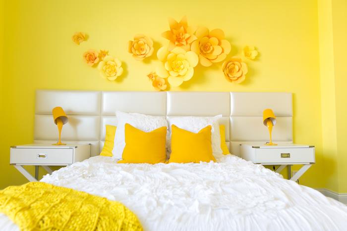 rumena barva spalnice za odrasle, belo in rumeno posteljnino, stenska dekoracija iz papirnate rože, bela nočna omarica