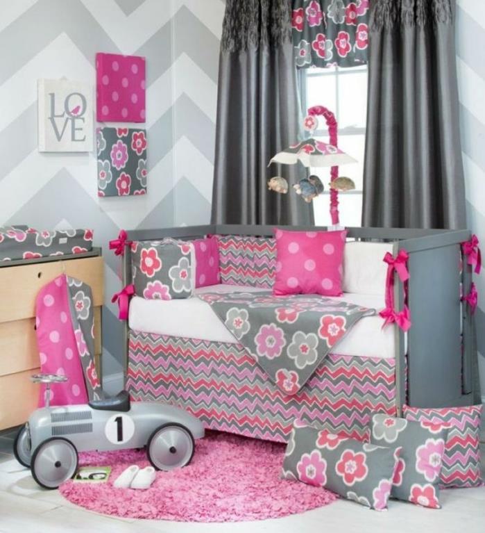 dekor spalnice za dekleta, sivo -bela stena, leseni predalnik, siva otroška posteljica, sive blazine in komplet postelj s sivimi in roza cvetličnimi vzorci, roza preproga, roza zavese