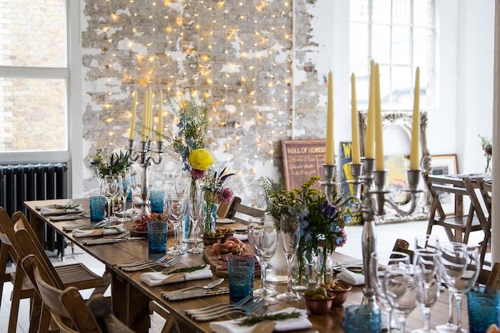 kaimiškas vestuvių dekoravimas, medinis stalas ir kėdės, mėlyni akiniai, stikliniai buteliai su gėlėmis, sidabrinis žvakidė su geltonomis žvakėmis, šviesi girlianda ant susidėvėjusios sienos
