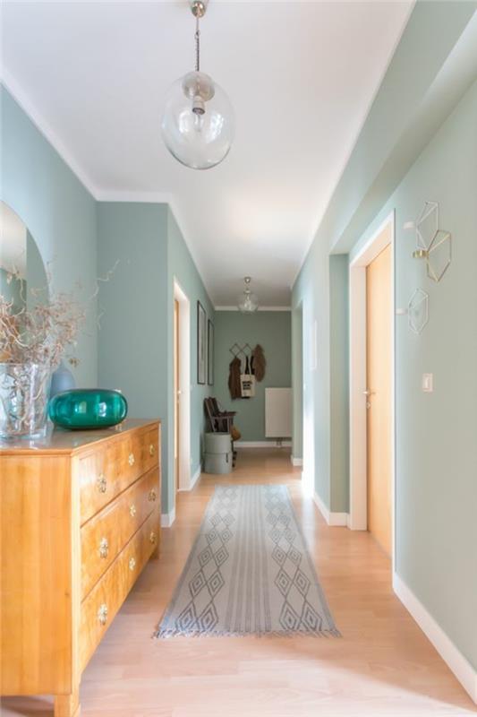pastelinės mėlynos ir baltos spalvos koridoriaus apdaila su šviesiai pilkos ir baltos spalvos kilimu, rutulio formos grindų lempa iš skaidraus stiklo, pastelinės mėlynos sienos, didelė šviesios medienos spintelė su dekoratyviniu dubeniu iš smaragdo žalio stiklo