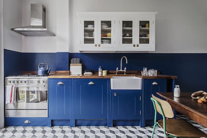 vintage kuhinjski dekor s polnočno modro kuhinjsko omarico in modro -belo stensko barvo bela visoka omara, sive in bele ploščice, miza iz surovega lesa