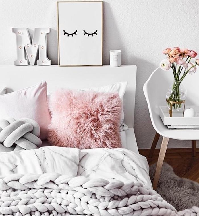 jauki rožinė pagalvėlė ir pilka lanko pagalvėlė, šviesiai pilka stambių tinklelių pledas, baltos sienos, dekoratyvinis galvūgalio rėmas, raidės žvakės LED žibintai, baltas skandinaviškas kėdės naktinis stalas