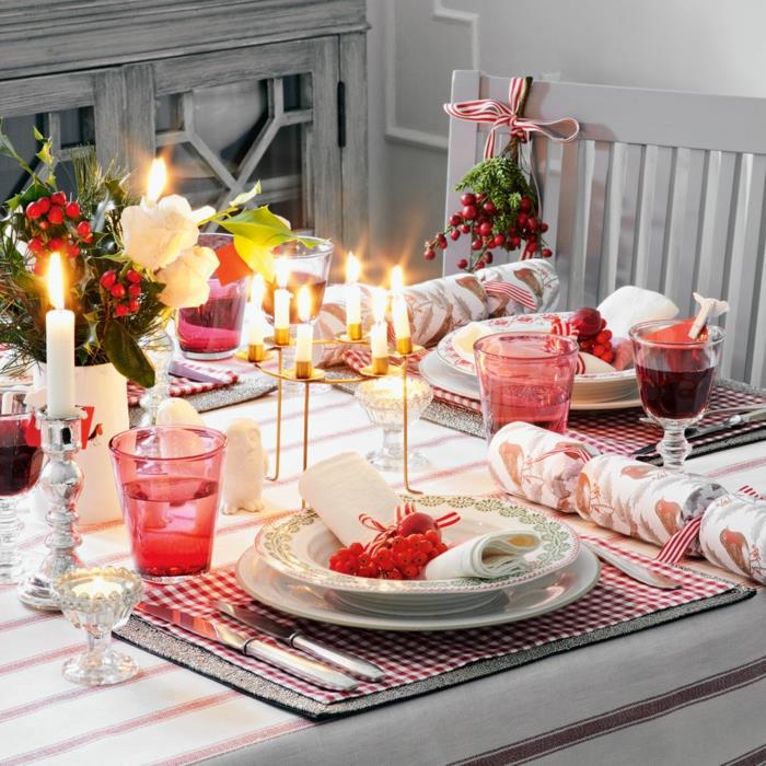 miza, okrašena za božič, pod kvadratnimi krožniki, prozornimi roza skodelicami, zlatim svečnikom, belimi svečami, svetlo sivim stolom in rdečimi jagodami