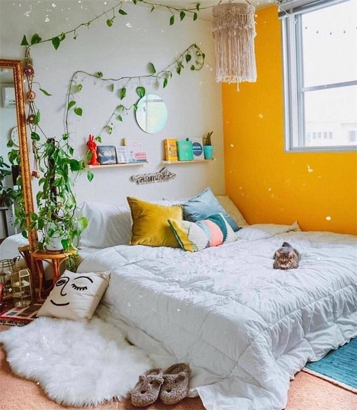 jauki atmosfera prašmatniame bohemiškame miegamajame, geltonais dažais akcentuojama siena, žema lova, papuošta spalvingomis pagalvėlėmis, žalias vijoklinis augalas interjere, kilimas iš avikailio, senovinis stačiakampis veidrodis
