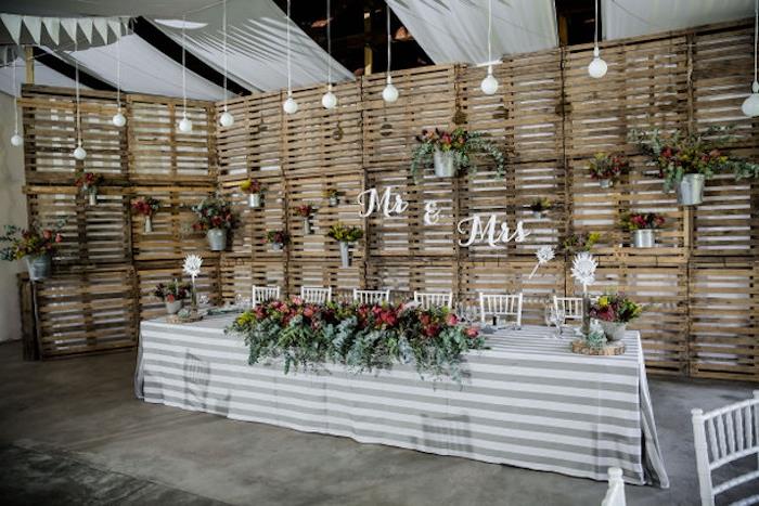 kmečka lesena stena iz palet z vedri in cvetličnimi pločevinkami, miza z belim in sivim prtom, cvetlični okras