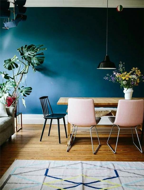 benzino mėlynas valgomojo dekoras, medinis stalas, rožinės ir juodos kėdės, medinės grindys, pilkas kilimas su geometriniais raštais, žalias augalas