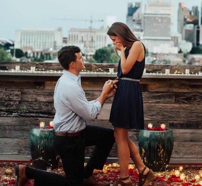 romantična poroka na strehi stavbe, ženska v črni obleki, moške kravatne hlače, tla pokrita z vrtnimi listi, svečami in svetlobnim vencem