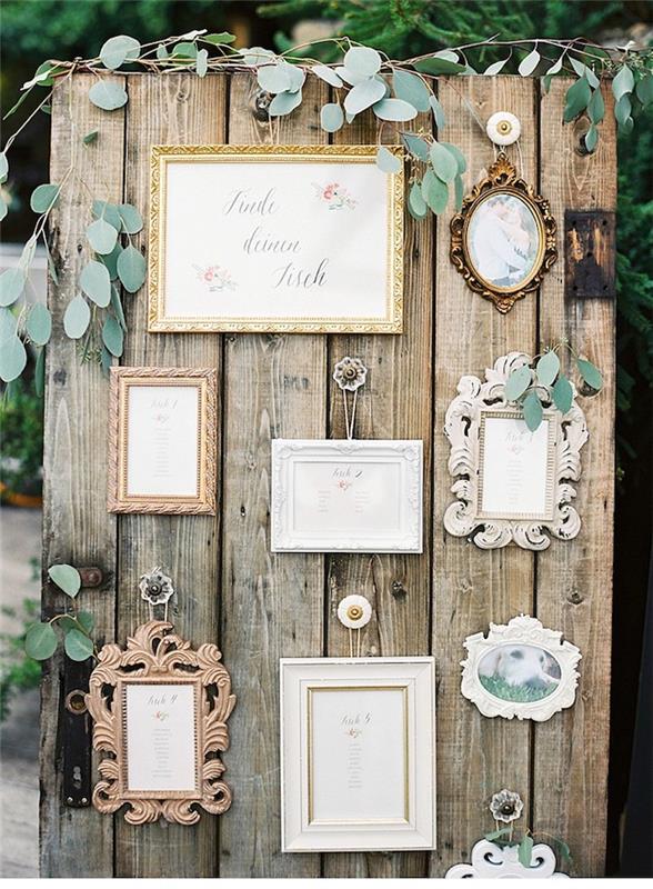 lauko vestuvių stalo planas, surinktos neapdorotos medienos lentos ir senoviniai nuotraukų rėmeliai su svečių vardais, maža žalia apdaila