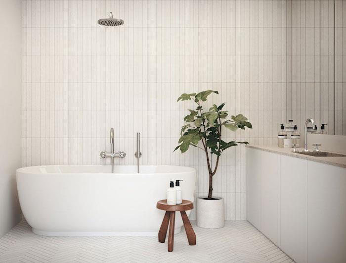 Kopalnica v skandinavskem slogu z belimi ploščicami na tleh in stenah ter ovalno samostoječo kadjo z lesenim stolčkom in zeleno rastlino