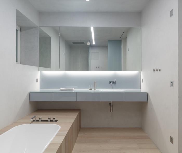 majhna kopalnica s kadjo in leseno talno oblogo z belimi stenami z velikim ogledalom in pod osvetlitvijo omare