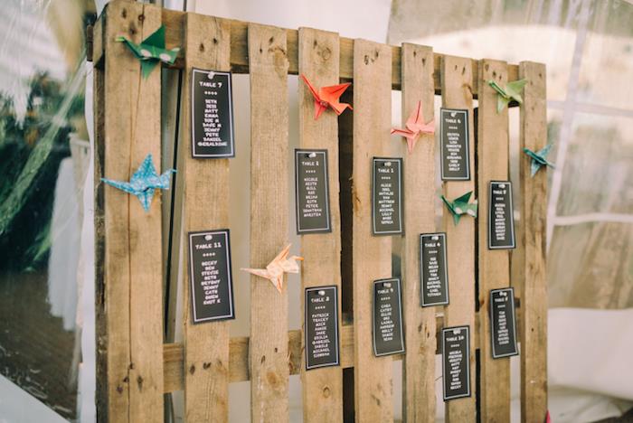 juodi svečių sąrašai, baltos raidės ant kaimiško medžio padėklo ir spalvingas origami popierius, originali vestuvių stalo plano idėja
