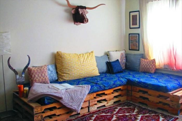 dekoratyvinis raudonos ir baltos spalvos kilimas, šalia kampinės padėklų sofos, dekoruotas mėlyno rašto audiniu ir keliomis skirtingomis pagalvėmis