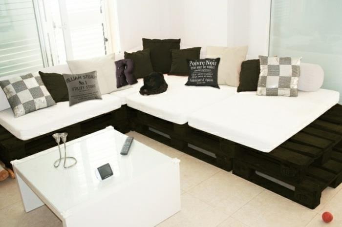 juodai balta kampinė sofa, su daugybe pagalvių pilkos, baltos ir juodos spalvos, skirtingų dydžių, su skirtingais raštais, baldai iš padėklų, modernus baltas stalas netoliese