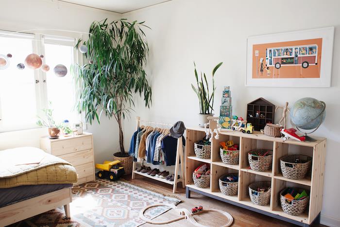 medinė žaislų laikymo lentyna su krepšeliais žaislams laikyti, baltos sienos, didelis žalias augalas, spalvingas kilimas, medinė lova, atvira vaikų drabužių spinta