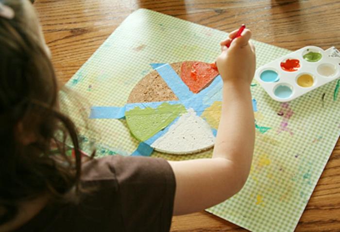 „Dyi“ dovanų idėjos, vaikas, dažantis kamštinį padėkliuką, uždėtą ant skirtingų spalvų veikiančio žaliai balto kilimėlio, paletė su mėlynais, raudonais, geltonais, baltais ir žaliais dažais ant medinio stalo