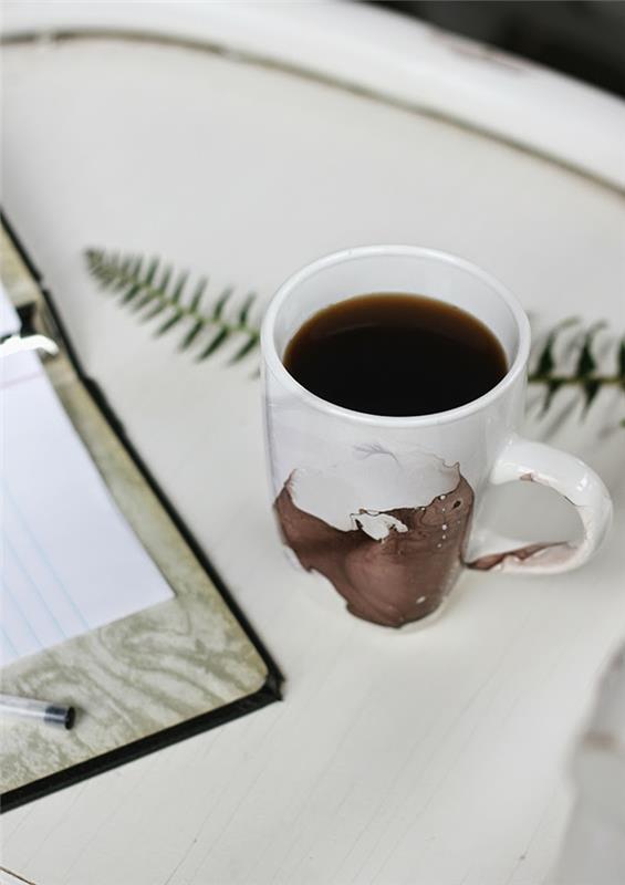 „pasidaryk pats“ dovanų idėjos, gražus dekoruotas puodelis kavos, padėtas ant balto stalo šalia atidarytos užrašų knygos