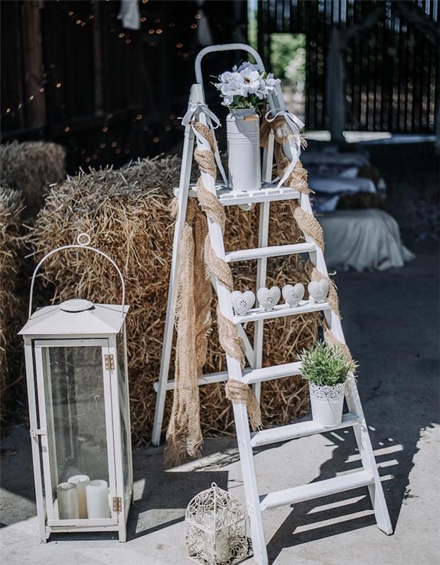 kaimiškas vestuvių dekoravimas, baltos dekoratyvinės kopėčios, baltos širdelės, gėlėtas pieno ąsotis ir didelis žibintas su žvakėmis viduje, šieno kupetos