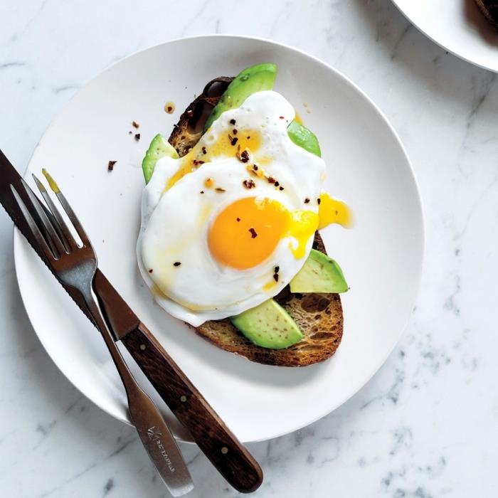 opečen kruh, z rezinami avokada, jajci na vrhu, uravnotežen obrok, na belem krožniku, nož in vilice na strani