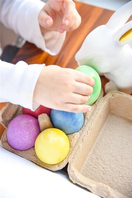 maža ranka, laikanti žalią, kiaušinio formos gleivių gabalėlį, virš kiaušinių dėžutės, kurioje yra raudonos ir mėlynos, violetinės ir geltonos spalvos kiaušinių, kaip padaryti gleives klijais