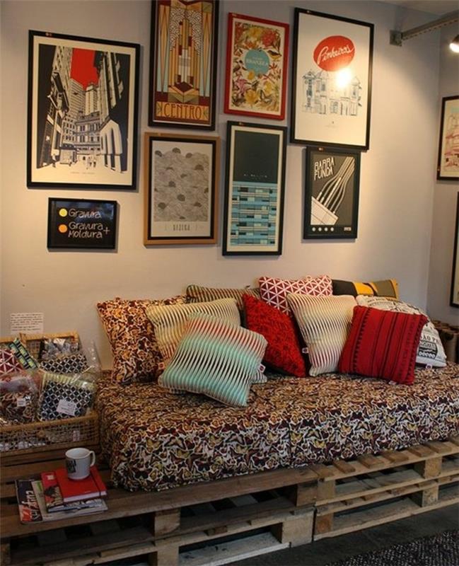 plakatai, dengiantys sieną, šalia padėklų sofos, čiužinys su įvairiaspalviu užvalkalu ir kelios pagalvėlės, skirtingų spalvų ir raštų