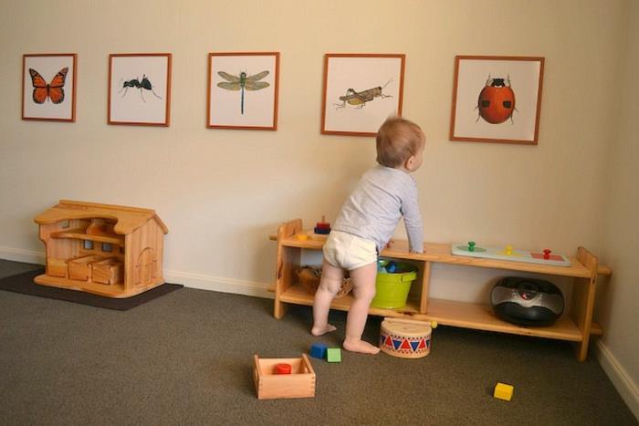 Montessori pedagogikos vaikų kambario dekoras, pilkas kilimas, medinė laikymo spintelė, medinis žaislų namelis, gyvūnų piešinių sienų dekoras