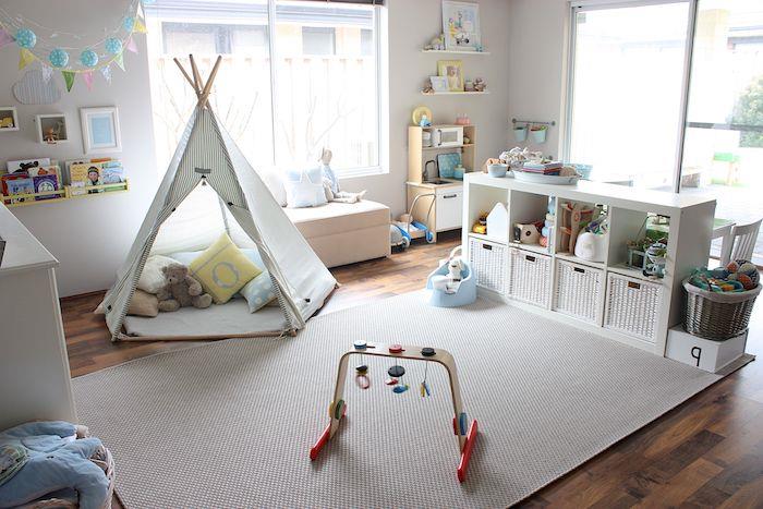 vaikiškas triukas su pagalvėlėmis ir žaisliukais viduje, pilkas žadinantis kilimas, žemai balta lentyna, rudas parketas, maža balta balta sofa prie lango