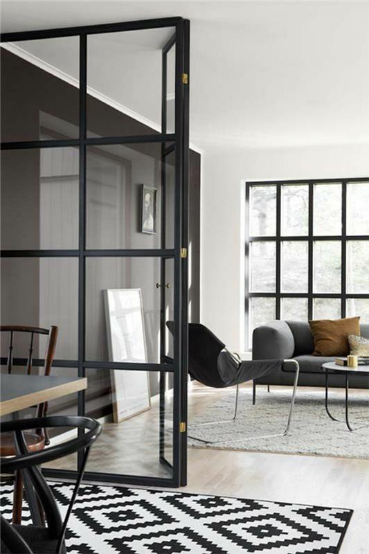 stiklo pertvara iš juodo metalo, su permatomais baltais langais, juodos ir baltos spalvos deimanto formos kilimas, lygus PVC parketas, sofa ir didelis pilkos spalvos fotelis