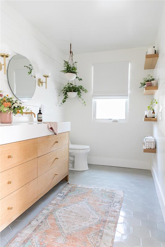 Zelene rastline okrasijo majhno kopalnico, sodobno kopalnico, okroglo ogledalo, omarico z lesenimi omarami in belim marmorjem za umivalnik