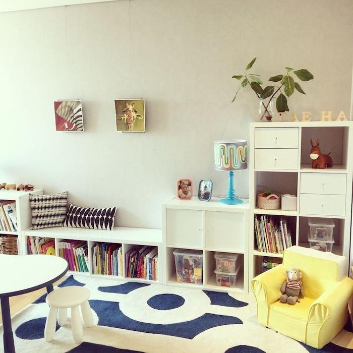 pilkos sienos, ilga žema lentyna knygoms ir žaislų dėžėms laikyti, vaikų rėmelių ir suoliuko sienų apdaila, baltas ir tamsiai mėlynas kilimas, žemas geltonas fotelis, žemas stalas ir kėdė