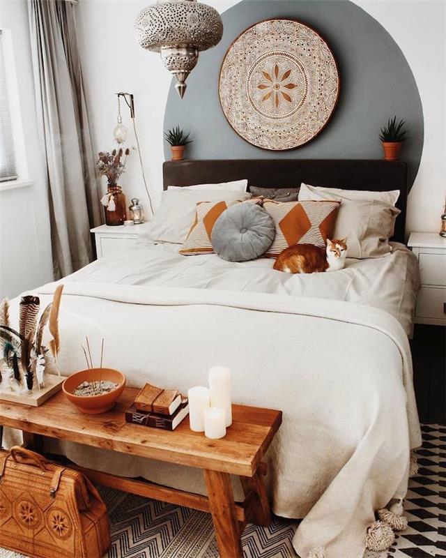 Berberų dekoras suaugusiųjų miegamajame, balta patalynė, rytietiškos pagalvėlės, medinis lovos galas, juodai baltas kilimas, rytietiška pakabinama lempa, geometrinė sienų tapyba
