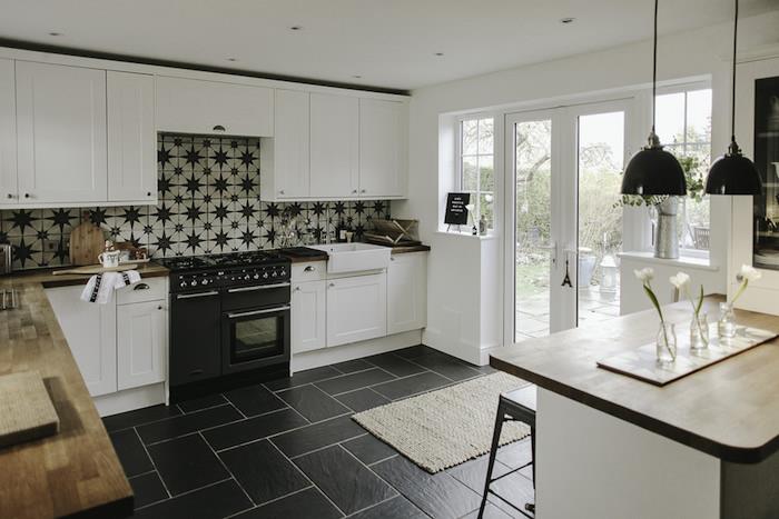baltos ir juodos virtuvės modelis su juodomis plytelėmis išklotomis grindimis, balti žemi ir aukšti baldai, baltos plytelių grindys su žvaigždutėmis, medinis stalviršis, centrinė sala su juodomis pakabomis