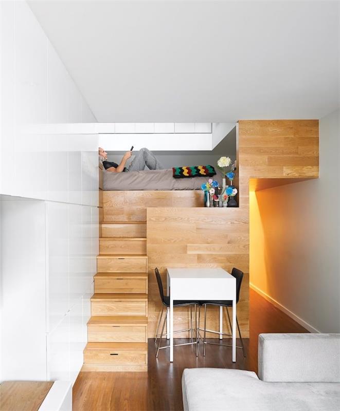 minimalistinis dizainas mažoje medinėje studijoje su lova virš virtuvės, baltu stalu ir juodomis kėdėmis, šviesiai pilka sofa