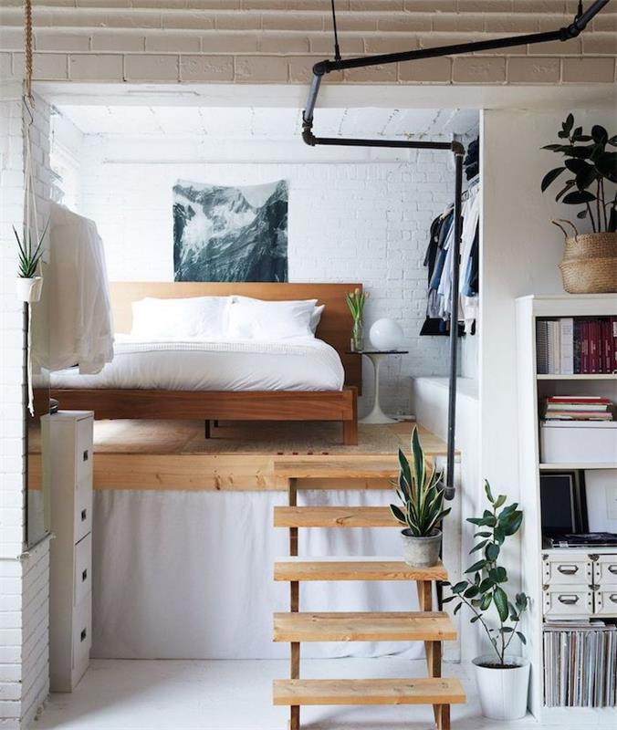 medinė lova, sumontuota ant medinės platformos su mažais mediniais laiptais, balinta plytų siena, atvira spinta, erdvę taupantis studijos dekoras