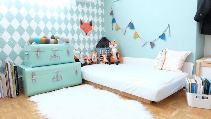 „Montessori“ miegamojo dekoras, medinė lova su baltu čiužiniu, dangaus mėlynos sienos, laikymo lagaminai, knygos ant grindų, lapės rašto deko