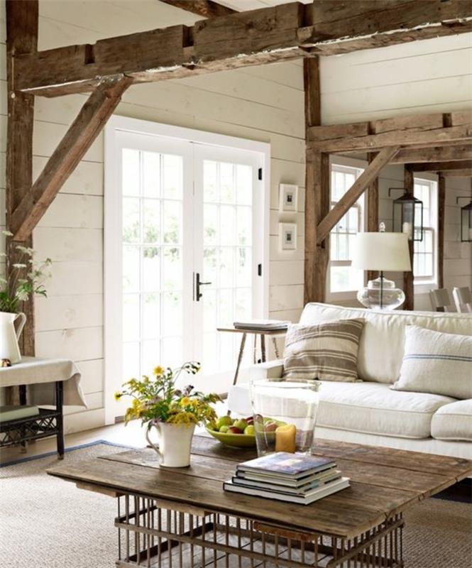 kaimiškas, prašmatnus kaimiškas dekoras, atvira sija, balta sofa, dekoratyvinės pagalvėlės, rudas kilimas, medinis lentų stalviršis ir metalinės dėžutės