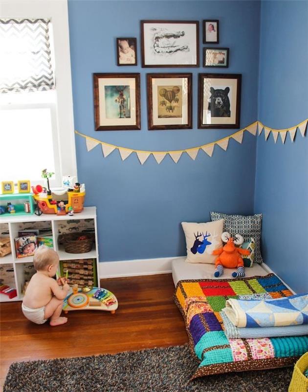 kūdikio berniuko kambario dekoras mėlynos spalvos su Montessori atmosfera, tamsiai mėlynos sienos, grindų čiužinys su spalvinga antklode, žemas žaislų laikymo įtaisas, siena dekoruota rėmeliais
