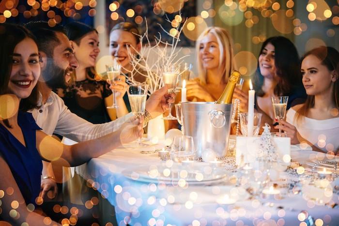Idejni večer za novo leto s prijatelji za mizo, okrašeno za novo leto, šampanjec, beli prti in krožniki, figurica božičnega drevesca, okras iz girlande iz srebrnega bisera