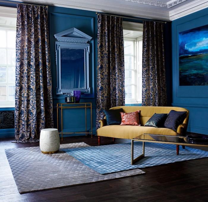rumen kavč, okrašen z blazinami v temno modri dnevni sobi s sivo preprogo in modro preprogo na parketu iz temnega lesa, sivimi in modrimi zavesami