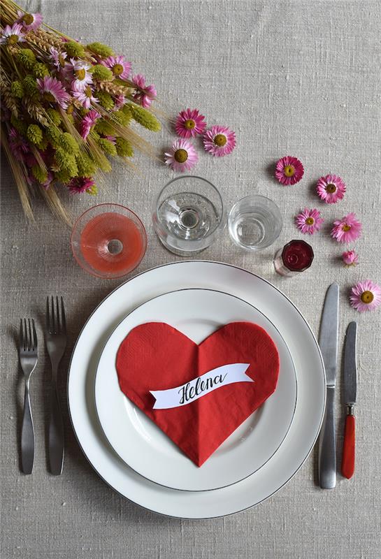 originali Valentino dienos stalo puošmena su sulankstoma raudona širdies servetėle ant baltos plokštelės ant pilkos staltiesės ir gėlių stalo dekoracija