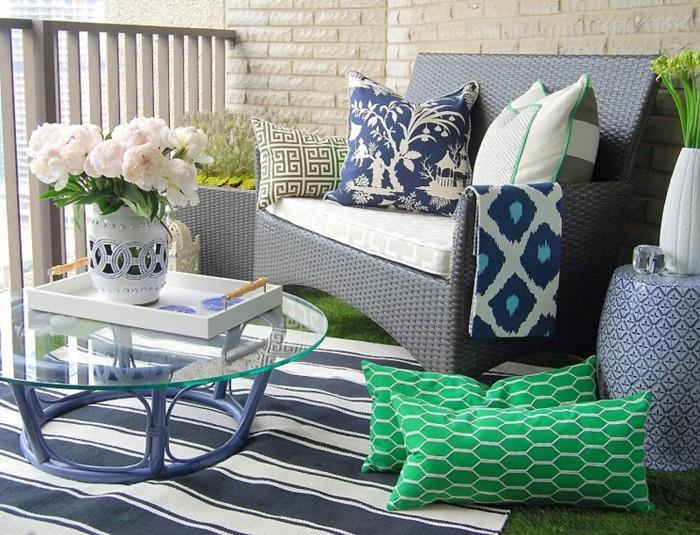balkonsko pohištvo iz sivega ratana, naslanjač, ​​okrašen z belimi, modrimi in zelenimi blazinami, modro -bela črtasta preproga, mizica s stekleno ploščo, rože v vazi