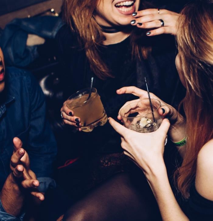 Besišypsanti mergina su kokteiliu rankoje, dvi merginos ir vyras, kalbantis vakarėlyje