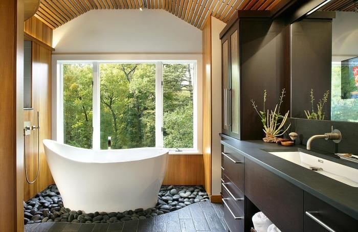 Azijietiškas vonios kambario išdėstymas su maža atskirai stovinčia vonia sode su pilkais akmenukais, vonios kambario apdaila pilka balta ir medinė