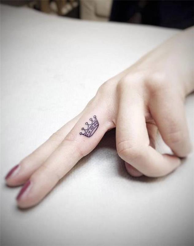 tetovaža majhne krone, tetovaža z prstanom, tetovaža s prsti, rdeč lak za nohte, roka počiva na belem pultu