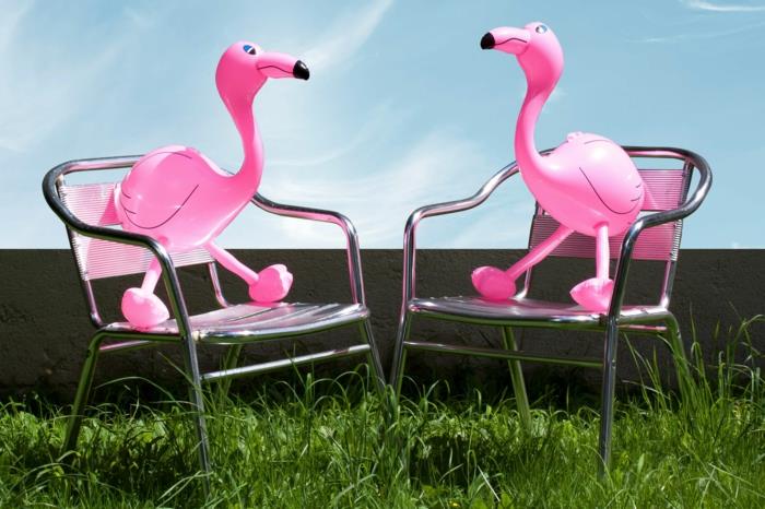 dva napihljiva flaminga, ki sedijo na kovinskih stolih srebrne barve v zeleni travi na vrtu, tropski deco na vrtu