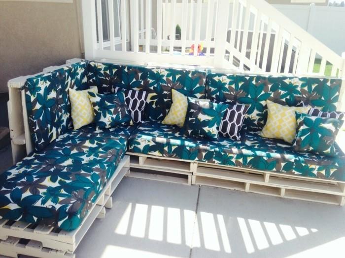 veranda prie baltų laiptų su didele kampine sofa, pagaminta iš šviesiai medinių padėklų, putų čiužiniais mėlynos, pilkos ir baltos gėlių raštu