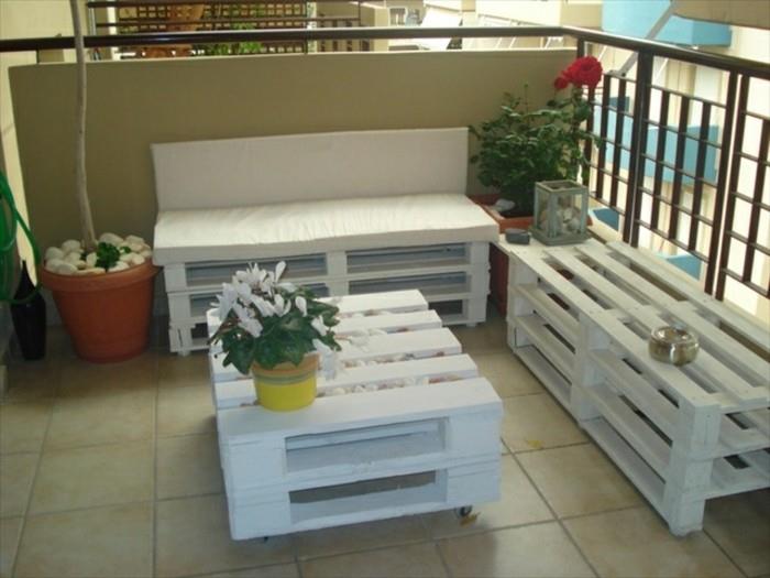 šviesiai smėlio spalvos plytelėmis dengtas balkonas, kuriame yra padėklų lauko baldai baltos spalvos, sofa ir suoliukas, kavos staliukas su gėlių vazonu