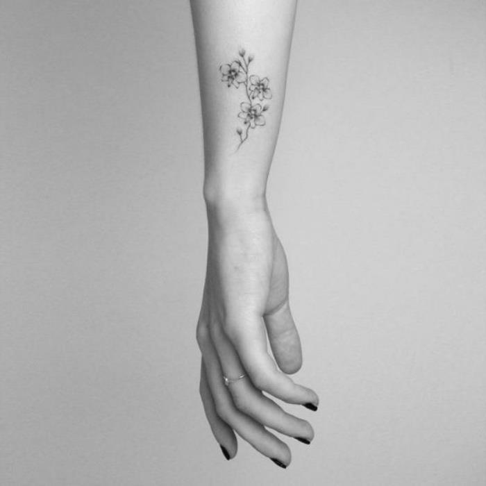 gėlės riešo tatuiruotė, moters ranka su juodu nagų laku, priešais baltą foną, mažos tatuiruotės su prasme